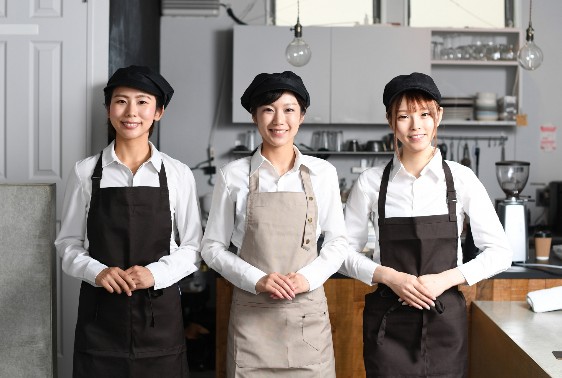 白いシャツに濃い色のエプロン、黒い帽子を身につけた3人が、モダンなカフェやレストランの厨房で並んで立ち、カメラに向かって微笑んでいる。背景には棚や食器、調理器具などが写っている。.