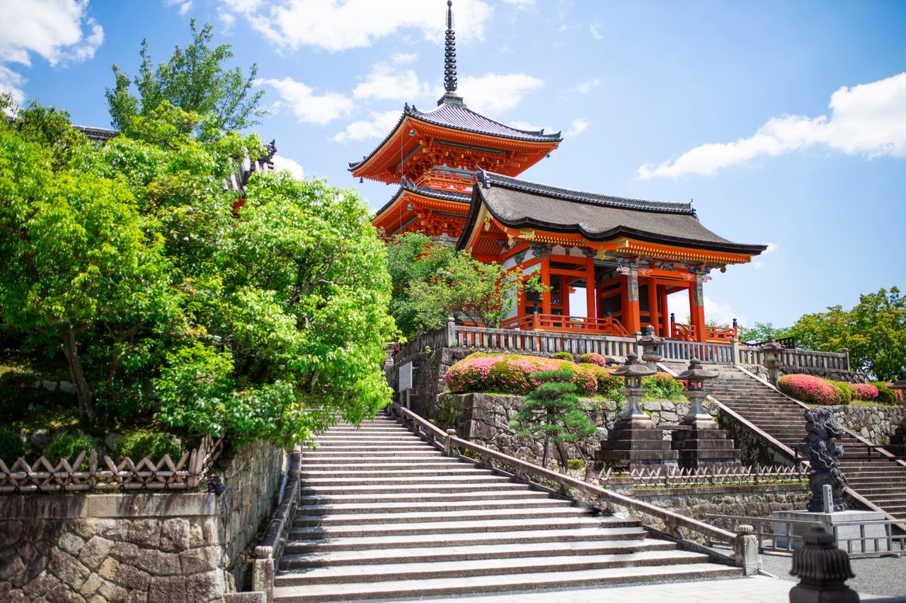 石段の上には、重層屋根の伝統的な日本の寺が佇み、青空に浮かぶ雲の下、緑の木々や手入れの行き届いた低木、色とりどりの花々に囲まれている。.