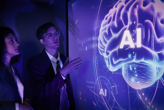 ビジネススーツ姿の2人が、中央に「AI」の文字が入った光り輝く脳のグラフィックが表示された大型デジタルスクリーンを見つめている。シーンは青と紫の色調で照らされ、未来的な雰囲気を醸し出している。.